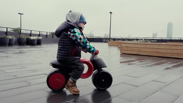 Side view of little kid in warm clothes learning to ride first toy run bike on wet after rain pavement on cloudy day