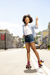 I love roller-skating. Happy curly girl smiling widely and showing V sign while roller-skating in the city center