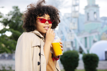 Fototapeta premium Favorite juice. Pleasant curly woman in a trench coat sipping orange juice through a straw while having a walk down the streets