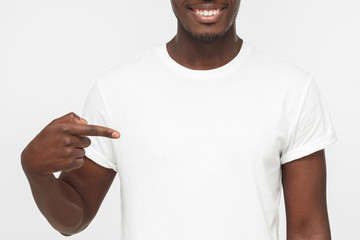 Smiling nice african american man pointing at his blank white t shirt with index finger, copy space...