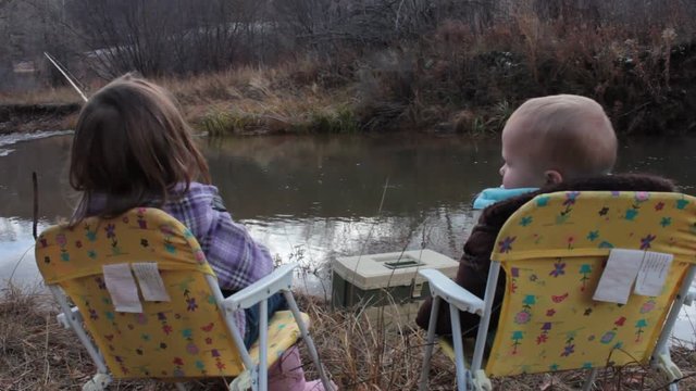 Two kids sitting in chairs by the river fishing. Video is taken from behind the kids, a river, tackle box, fishing pole, and trees can be seen.