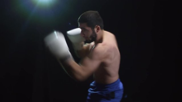 Medium shot of bearded bare chested male boxer in gloves throwing punches isolated on black background with studio light shining from above