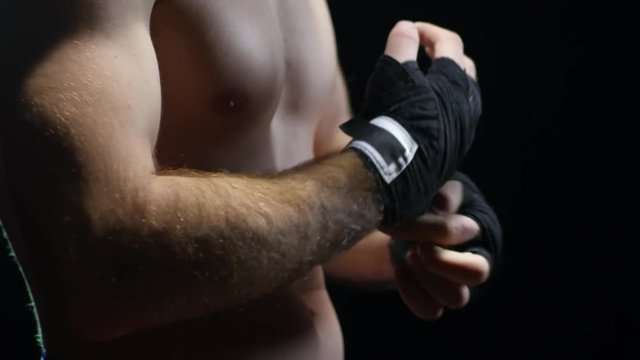 Studio shot with PAN of unrecognizable muscular male fighter putting on wrist wraps and warming up hands before fight