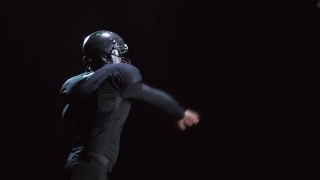 Studio shot with side view of male American football player in protective helmet and full uniform throwing ball and running isolated on black background