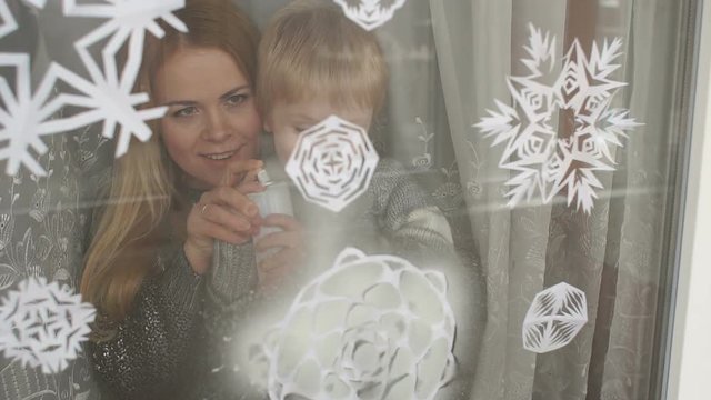 Mom and her son decorate the windows for Christmas with snowflakes and artificial snow, they spray artificial snow on paper snowflakes on the window.