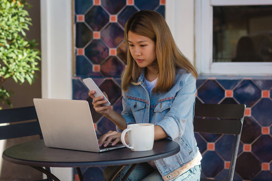 Cheerful asian young woman sitting in cafe drinking coffee and using smartphone for talking, reading and texting. Attractive asian woman holding a cup of coffee while working on laptop.