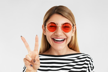 People, joy, happiness, success and positive emotions concept. Picture of happy overjoyed European hipster girl wearing round shaped shades with pink lenses, laughing and making peace gesture