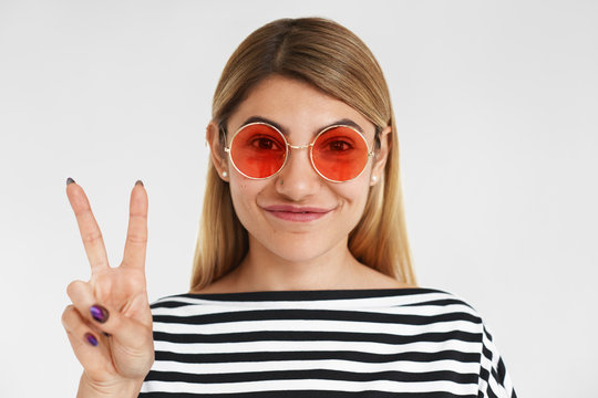Beauty, style, glamour and fashion concept. Charming stylish young blonde female wearing striped top and trendy pink sunglasses in round shape smiling happily, showing peace sign at camera