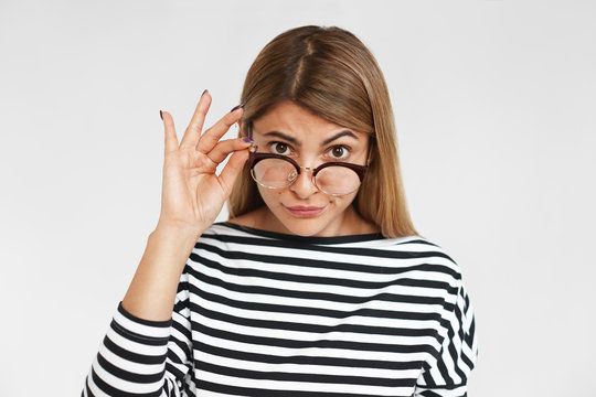 People, style, fashion, eyewear and optics concept. Stern beautiful young lady wearing striped top, lowering her stylish eyeglasses in black frame and straring at camera with scrutinizing look