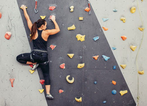 Female fitness professional climber training at bouldering gym. Muscular  woman with athletic body dressed in black, climbing on artificial colourful  rock wall. Active lifestyle and bouldering concept. Stock Photo