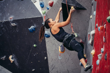 Professional climber woman in black outfit, reaching the top of artificial bouldering wall while practicing in bouldering gym. Low angle vertical view of stretching on air muscular female body.