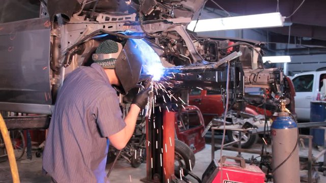 Worker welds the inner apron to a new frame rail on a wrecked car. Sparks fall down and around the welder, includes audio of mig welding.