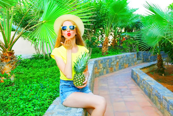 Summer mood! Fashion woman sends an air kiss holds a pineapple on a palm trees background