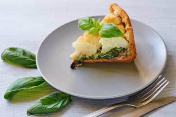 A piece of healthy cauliflower pie on the gray plate decorated with fresh basil leaves and vintage silver knife and fork. Vegetarian cauliflower tart on the gray background. Close up view