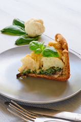 A piece of healthy cauliflower pie on the gray plate decorated with fresh basil leaves and vintage silver knife and fork. Vegetarian cauliflower tart on the gray background. Side view