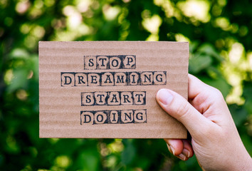 Woman hand holding cardboard card with words Stop Dreaming Start Doing against green nature background.