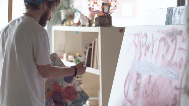 Focused man painting a picture at the easel, wearing white t-shirt and blue bandana as working in spacious light studio, concept of hard work to achieve a goal