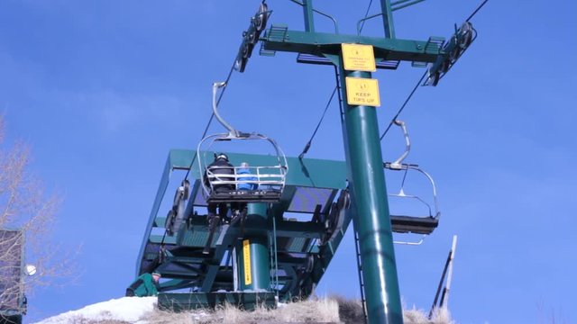 Skiers unload at the top of a hill. POV from under the lift looking up. a worker can be seen raking the snow.