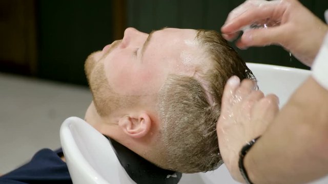 Preparing for the haircut. Male barber washing the head of the young man in barber's. 4K