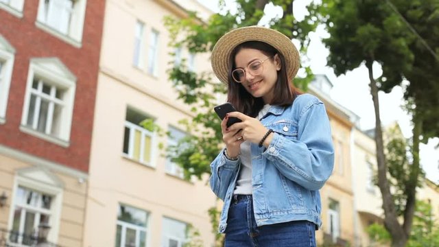 pretty business lady wearing jeans jacket and straw hat using smart phone cellphone while standing on the street spring female fashion look onlie social networking communicate chattig view from below