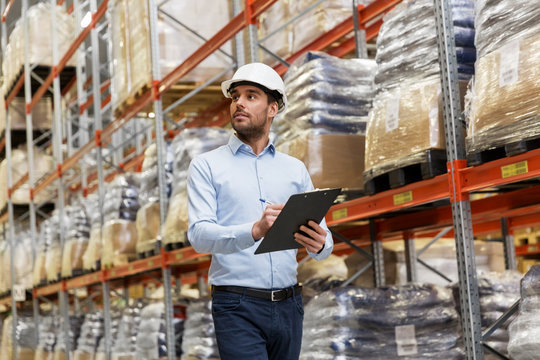 logistic business, shipment and people concept - businessman in helmet with clipboard checking goods at warehouse