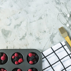 Raspberries and blackberries in metal form for cupcakes next to cooking tools on a gray table. Summer baking concept. Top view, flat lay