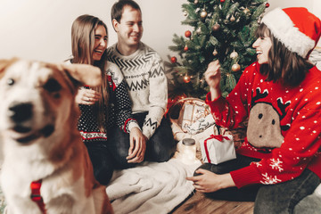 happy family in stylish sweaters and cute funny dog exchanging gifts at christmas tree with lights....