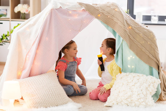 childhood, hygge and friendship concept - little girls with torch light having fun in kids tent or teepee at home
