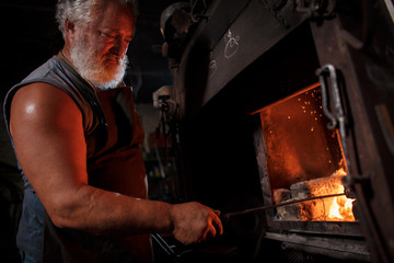 The smith artisan in his apron heats the workpiece in the burning furnace in the smith