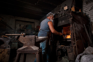 The smith artisan in his apron heats the workpiece in the burning furnace in the smith