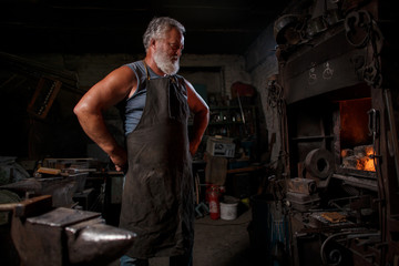The smith artisan in his apron heats the workpiece in the burning furnace in the smith