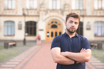 Portrait of a man with a beard and a blue T-shirt stands against the background of the building and looks at the camera. Student handsome man posing against a background of university buildings.