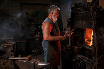 The smith artisan in his apron heats the workpiece in the burning furnace in the smith