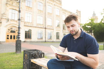 Portrait of a beautiful man with a beard and a black T-shirt sitting on a bench on the background of architecture and reading a book. Student reads a book at a university campus.