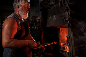 The smith artisan in his apron heats the workpiece in the burning furnace in the smith