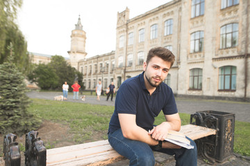 Handsome man sitting on a bench at a university campus with books and looking at the camera. Portrait of a man with a beard on a bench with books in his hands on the background of the architecture