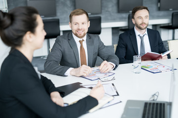 Group of three modern entrepreneurs sitting at meeting table in conference room and smiling while discussing startup project, copy space
