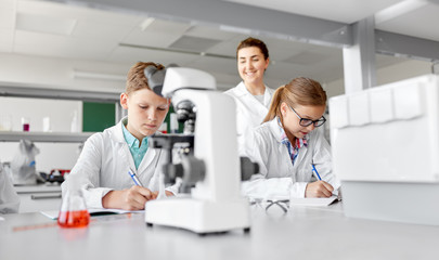 education, science and children concept - teacher and students studying chemistry at school laboratory and writing to workbooks