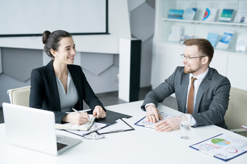 Fototapeta na wymiar Portrait of two young entrepreneurs sitting at meeting table in conference room, focus on smiling businesswoman listening to colleague, copy space