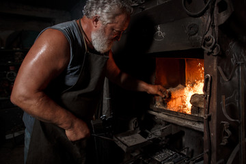 The smith artisan in his apron heats the workpiece in the burning furnace in the smith