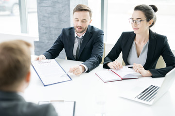 Portrait of two successful business people, man and woman,  presenting contract to partner sitting across meeting table in conference room, copy space