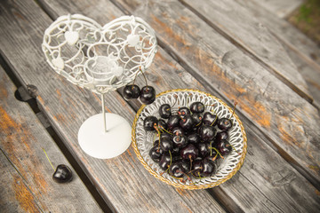 fresh red cherries berries in a white plate on the wooden table with candlestick in the shape of the heart outdoors in the garden. Romantic place.