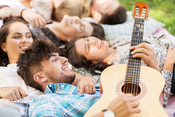 Fototapeta premium friendship, leisure and summer concept - group of happy smiling friends playing guitar and chilling on picnic blanket