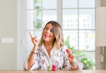 Young woman at home eating macarons very happy pointing with hand and finger to the side