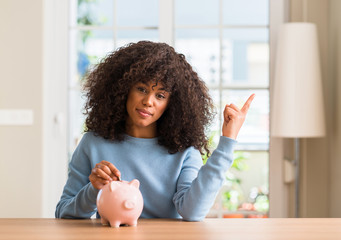 African american woman saves money in piggy bank very happy pointing with hand and finger to the...
