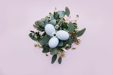 Beautiful and tender easter setup: three light blue easter eggs in a nest made of eucalyptus leaves, flowers and branches on the tender pastel pink background, top view, flat lay 