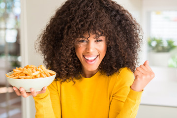 African american woman holding a plate with potato chips at home screaming proud and celebrating victory and success very excited, cheering emotion