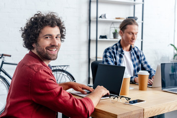 young man using laptop with blank screen and smiling at camera while working with colleague in office