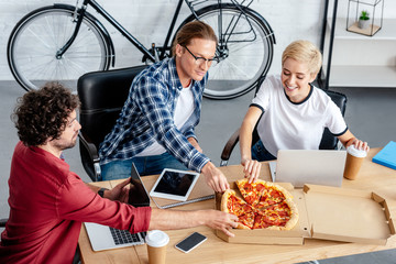 smiling young men eating pizza and using laptops in office