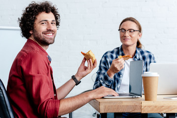 smiling young men eating pizza and using laptops in office